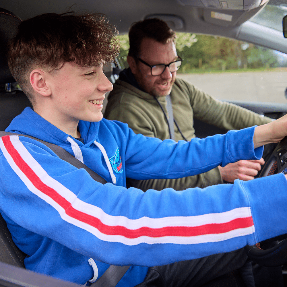 A teenage boy in a blue hoodie sits in the driver's seat of a car, smiling, while an adult man in glasses sits beside him, both wearing seatbelts.