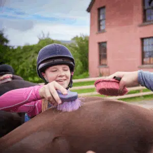 A girl in a helmet smiles while brushing a horse with a purple brush, with another person using a red brush nearby. A red building is visible in the background.