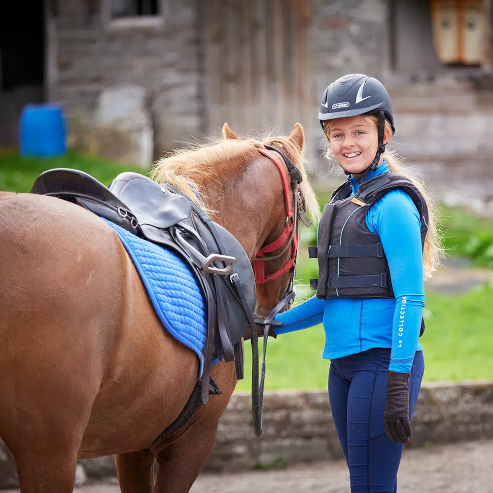 A young person in equestrian gear stands beside a saddled brown horse, holding the reins and smiling at the camera outdoors.