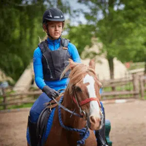 A person wearing a helmet and vest rides a brown horse with a red bridle in an outdoor riding arena with trees and fencing in the background.