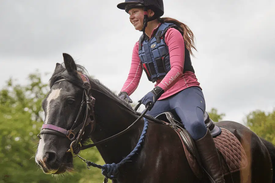 A woman wearing a helmet and riding gear rides a black horse outdoors, with greenery and a cloudy sky in the background.