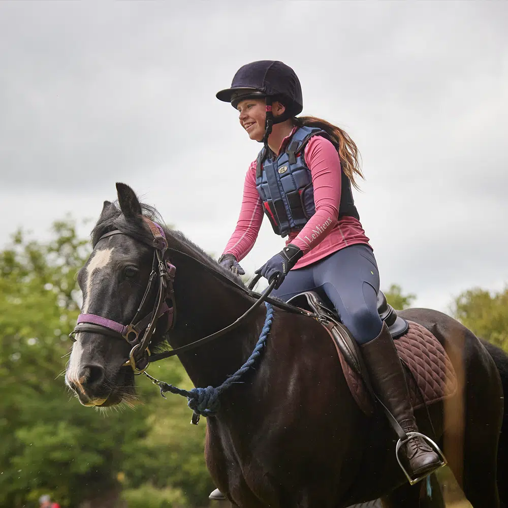 A woman wearing a helmet and riding gear rides a black horse outdoors, with greenery and a cloudy sky in the background.