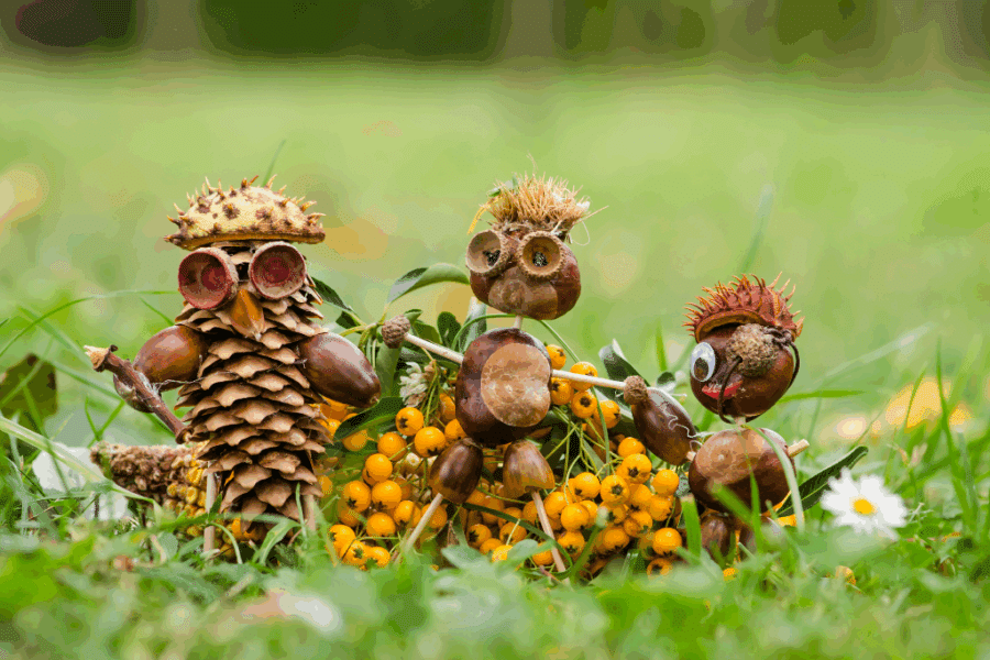 Three small figures made from pine cones, chestnuts, acorns, and natural materials are arranged on grass with yellow berries in the background.