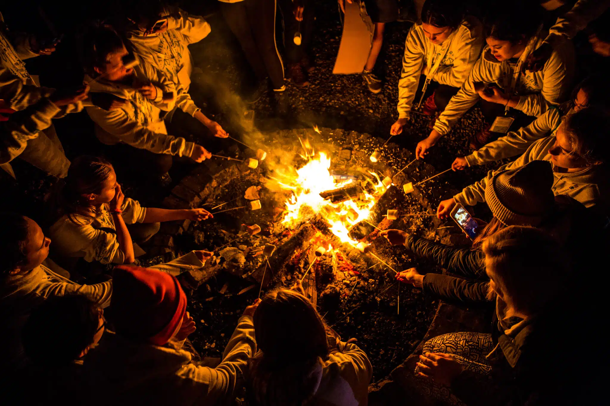 A group of people sits around a campfire at night, roasting marshmallows on sticks.