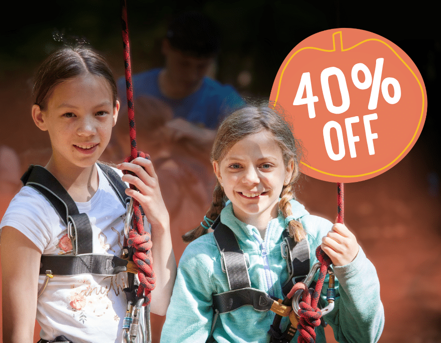 Two young girls wearing harnesses and holding ropes outdoors during an exciting PGL Adventure Holidays experience, with a graphic showing 