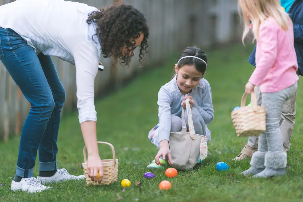 An adult and two children collect colorful Easter eggs in baskets on a grassy lawn.