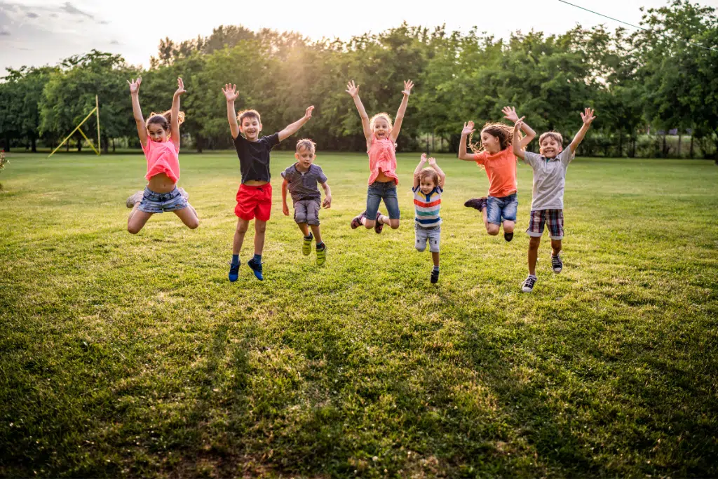 Seven children are jumping with arms raised on a grassy field, with trees and sunlight in the background.