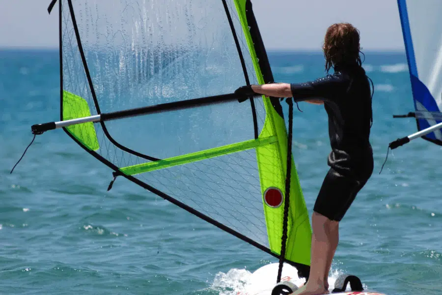 Person in a wetsuit windsurfing on the ocean, holding onto the sail and standing on the board with water and a clear sky in the background.