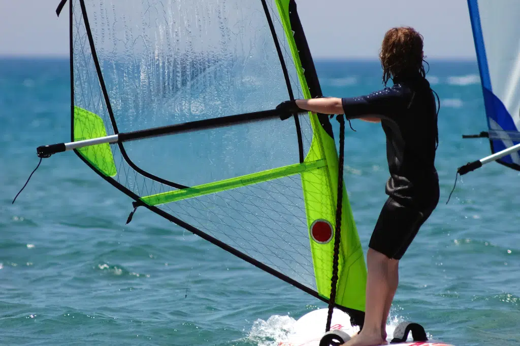 Person in a wetsuit windsurfing on the ocean, holding onto the sail and standing on the board with water and a clear sky in the background.