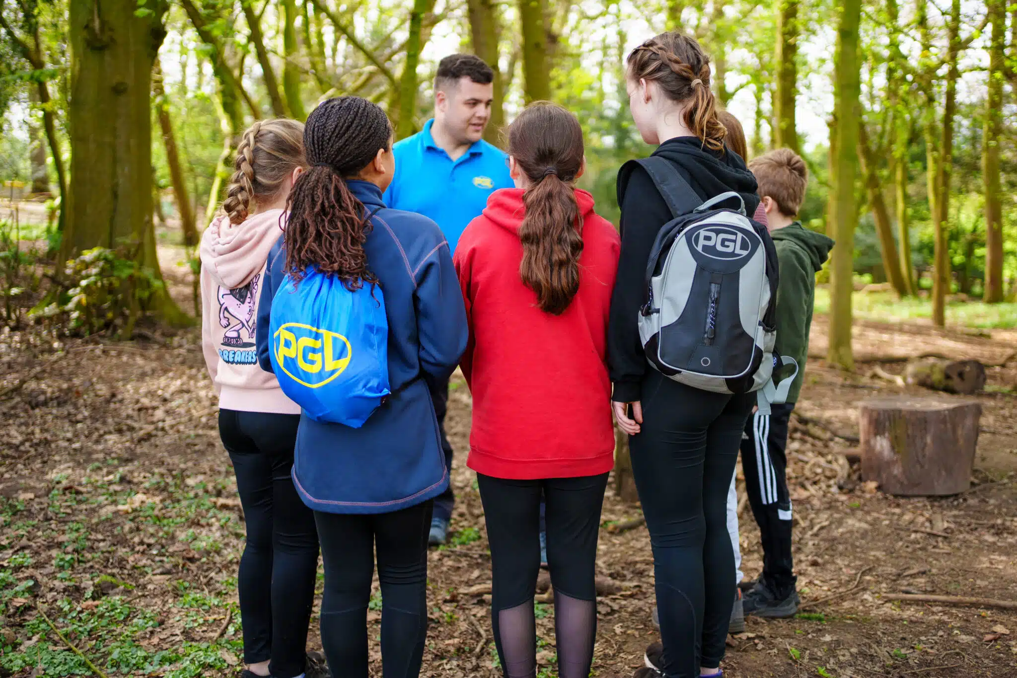 A group of children with backpacks listen to an instructor in a blue shirt during an outdoor activity in a wooded area.