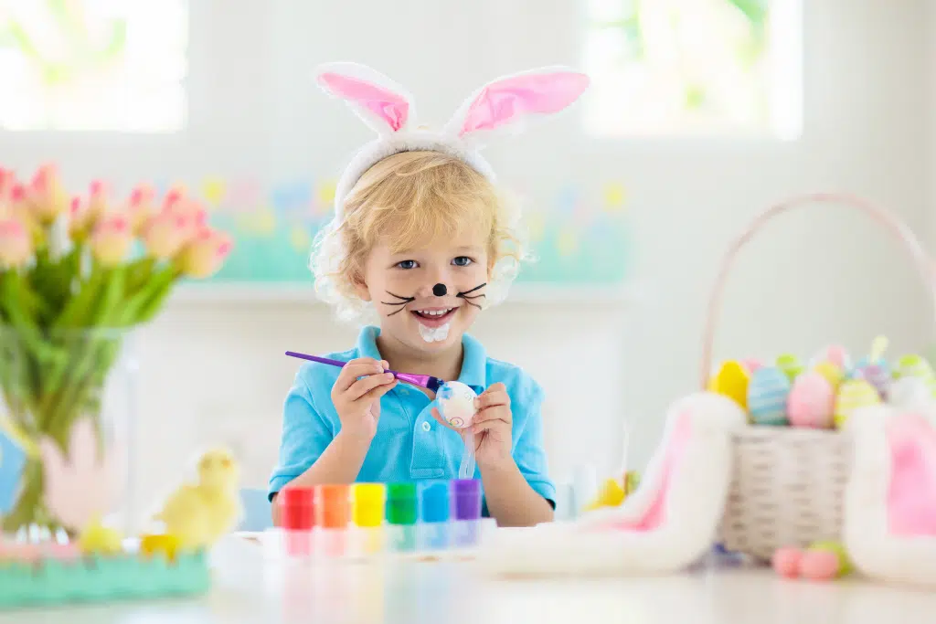 Young child wearing bunny ears and face paint sits at a table, painting an Easter egg with paint pots and brushes, surrounded by decorated eggs and spring flowers.
