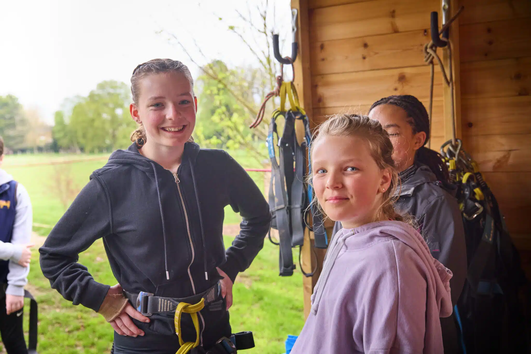 Two girls wearing harnesses stand in a wooden shed, smiling at the camera, with climbing equipment hanging on the wall behind them.