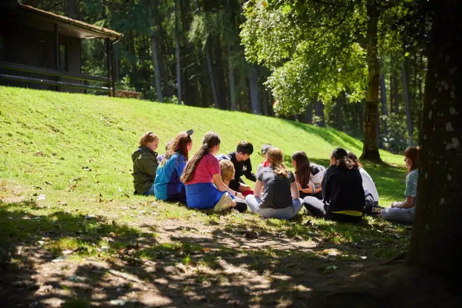 A group of people sit in a circle on the grass under trees near a cabin, engaged in conversation in a sunlit outdoor setting.