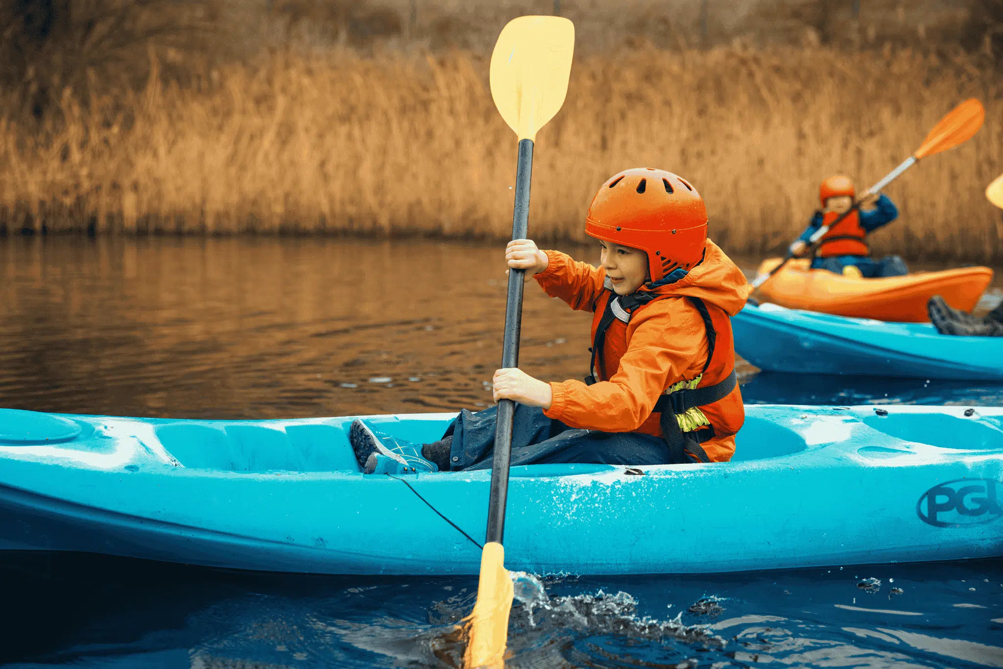 A child wearing a helmet and life jacket paddles a blue kayak on a calm body of water, with other children kayaking in the background.