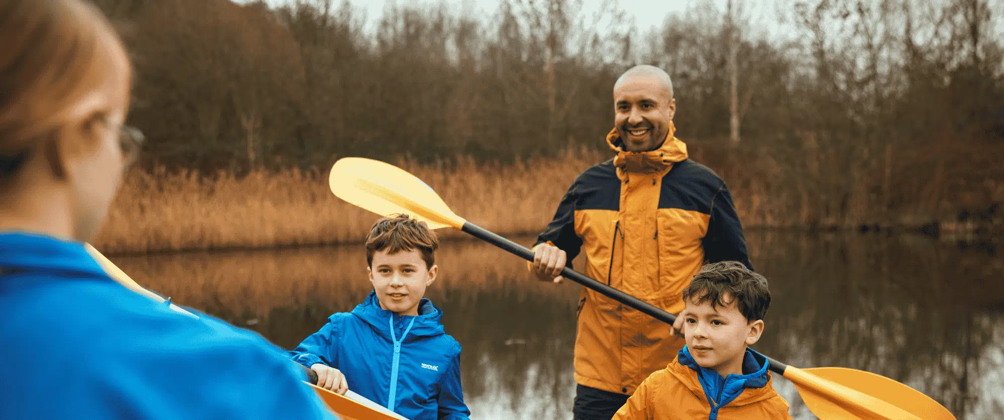 A man and two boys, all in jackets, stand by a lake with paddles, talking to a woman in the foreground. Trees and reeds are visible in the background.