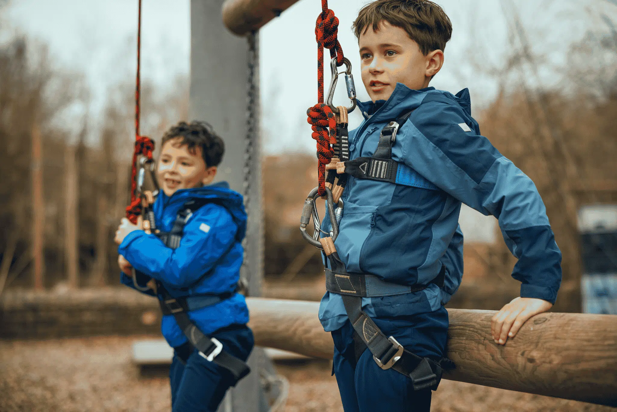 Two boys in blue jackets wearing safety harnesses prepare for a zip wire or ropes course outdoors, holding onto ropes and standing by a wooden rail.