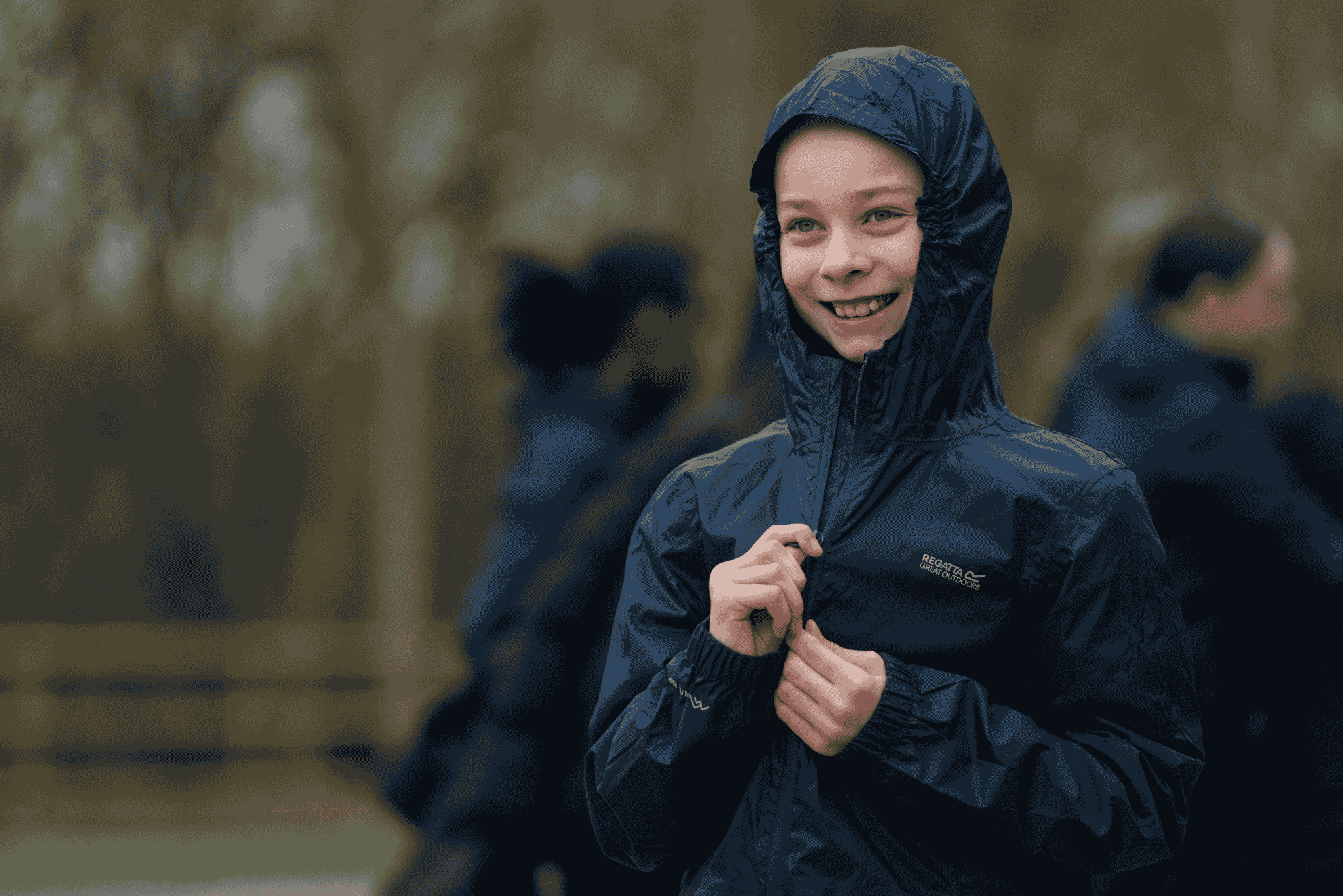 A person wearing a hooded navy raincoat is zipping it up and smiling outdoors, with blurred people and trees in the background.