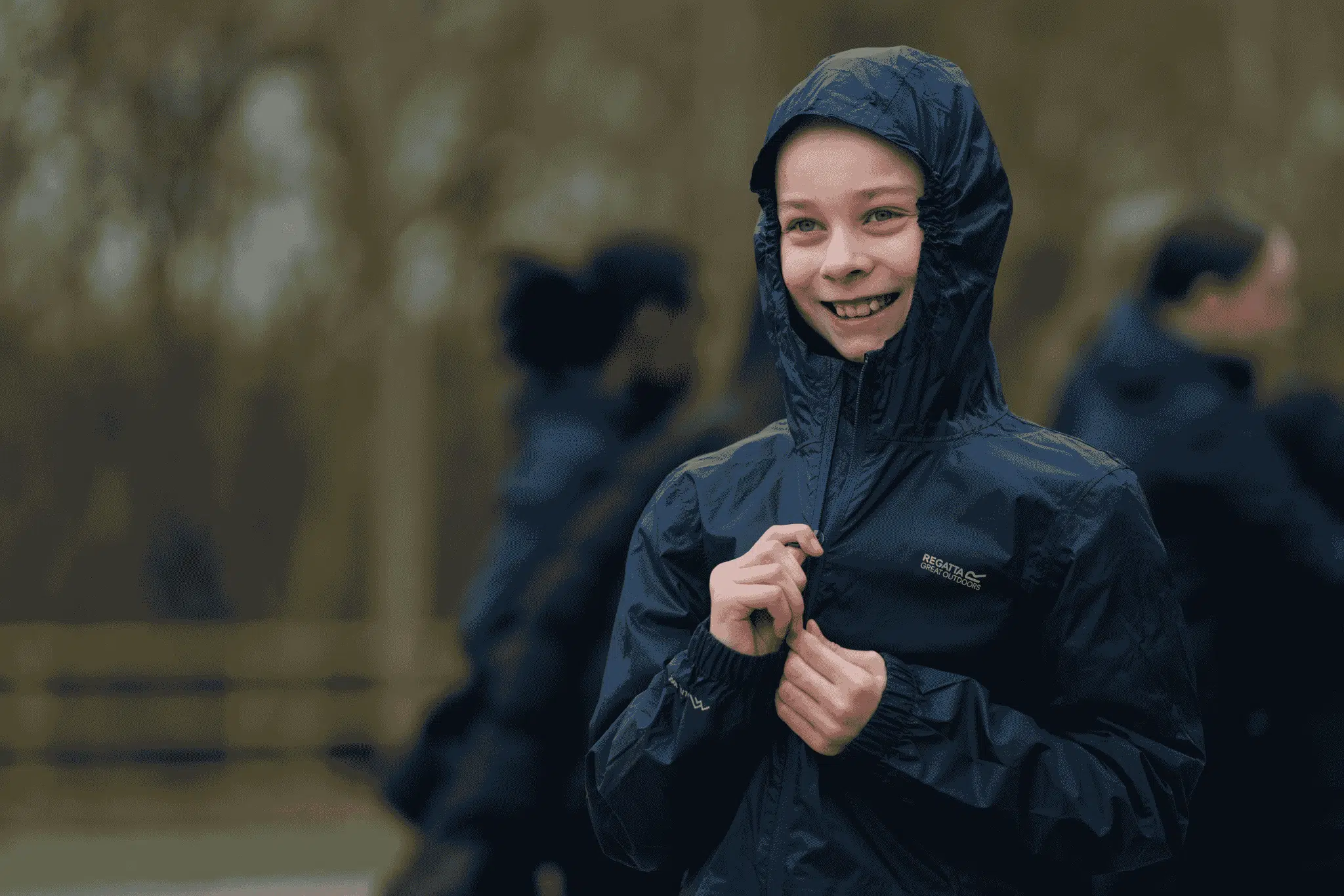 A person wearing a hooded navy raincoat is zipping it up and smiling outdoors, with blurred people and trees in the background.
