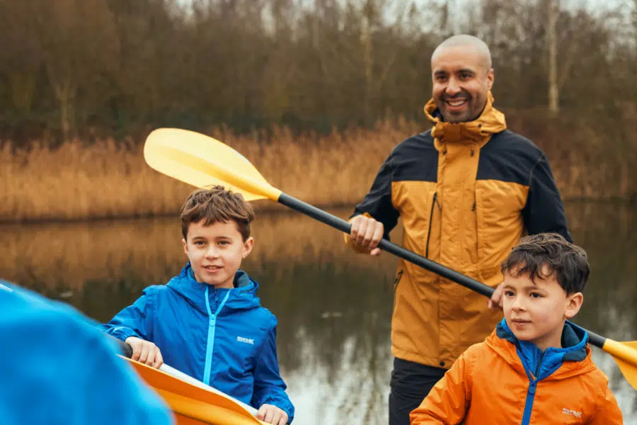 An adult and two children with paddles stand by a lake, dressed in outdoor jackets, with trees and reeds in the background.