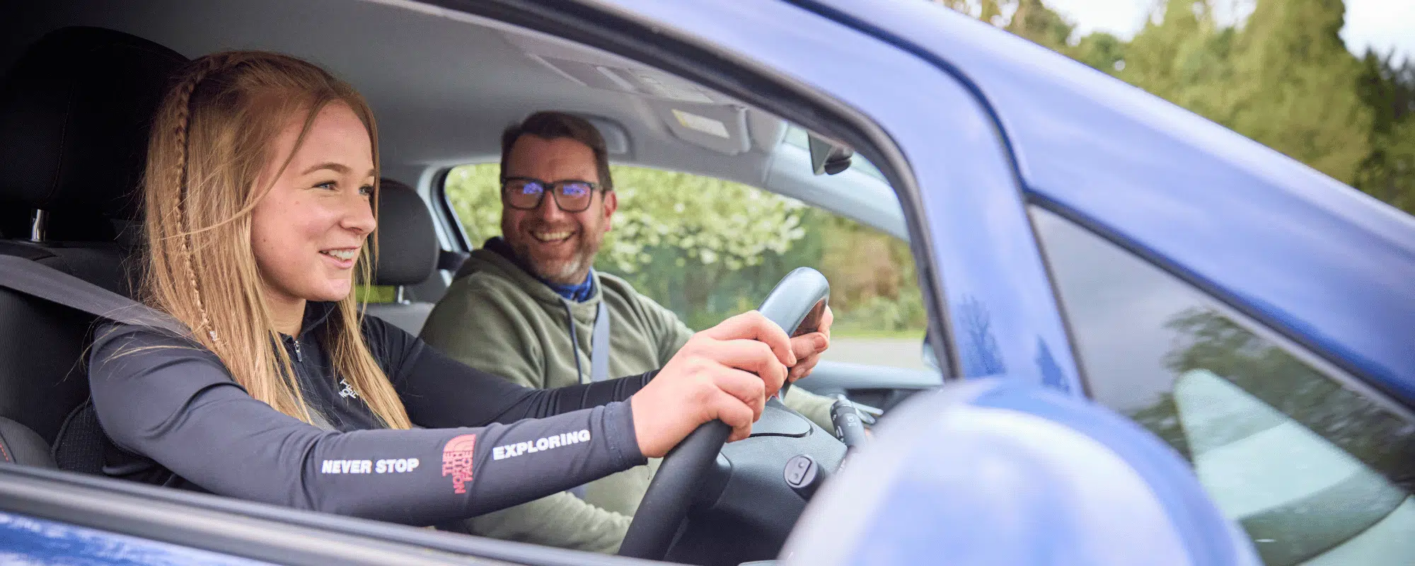 A young woman drives a car while a man in the passenger seat smiles; both appear engaged and happy, perhaps en route to a PGL Adventure Holidays destination, with trees visible outside the vehicle.