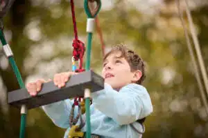 A boy wearing a light blue jumper holds onto a rope obstacle, looking up with determination whilst enjoying an outdoor climbing activity on his PGL Adventure Holiday.