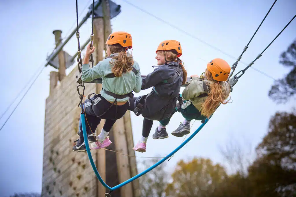 Three children wearing helmets and harnesses sit on a swing suspended outdoors during PGL Adventure Holidays, near a wooden climbing wall, with trees visible in the background.