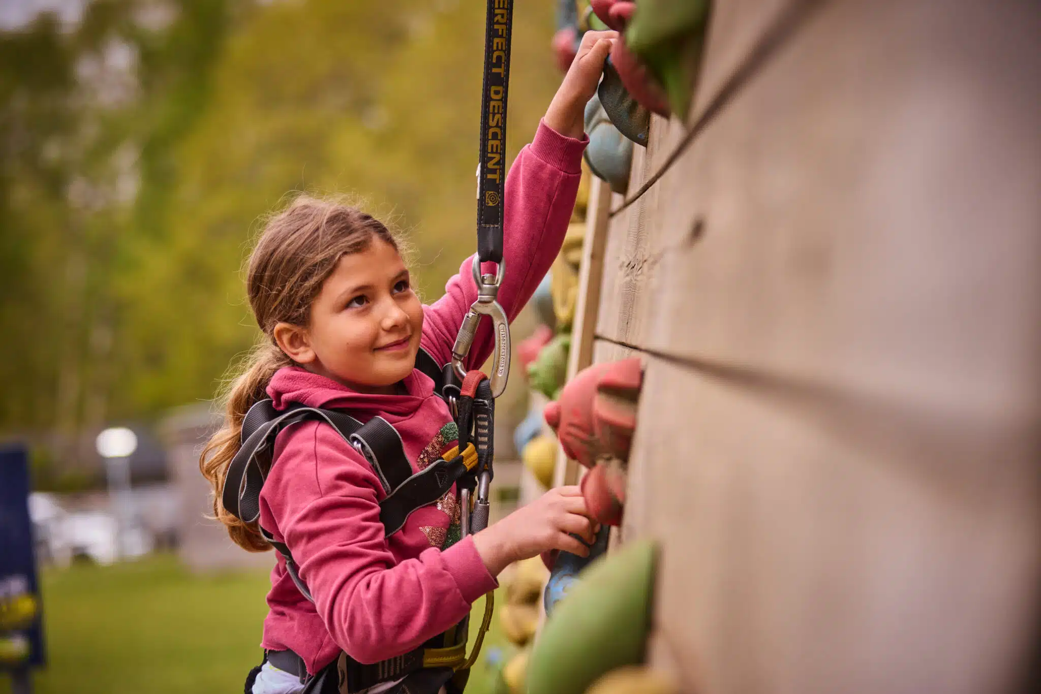 A young girl wearing a harness and safety rope climbs an outdoor wall at PGL Adventure Holidays, gripping colourful handholds.