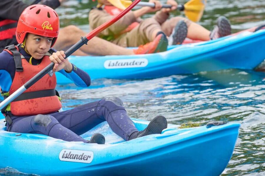 Several people in kayaks paddle on a body of water. The child in the foreground is in a blue kayak and wearing a red helmet and life vest.