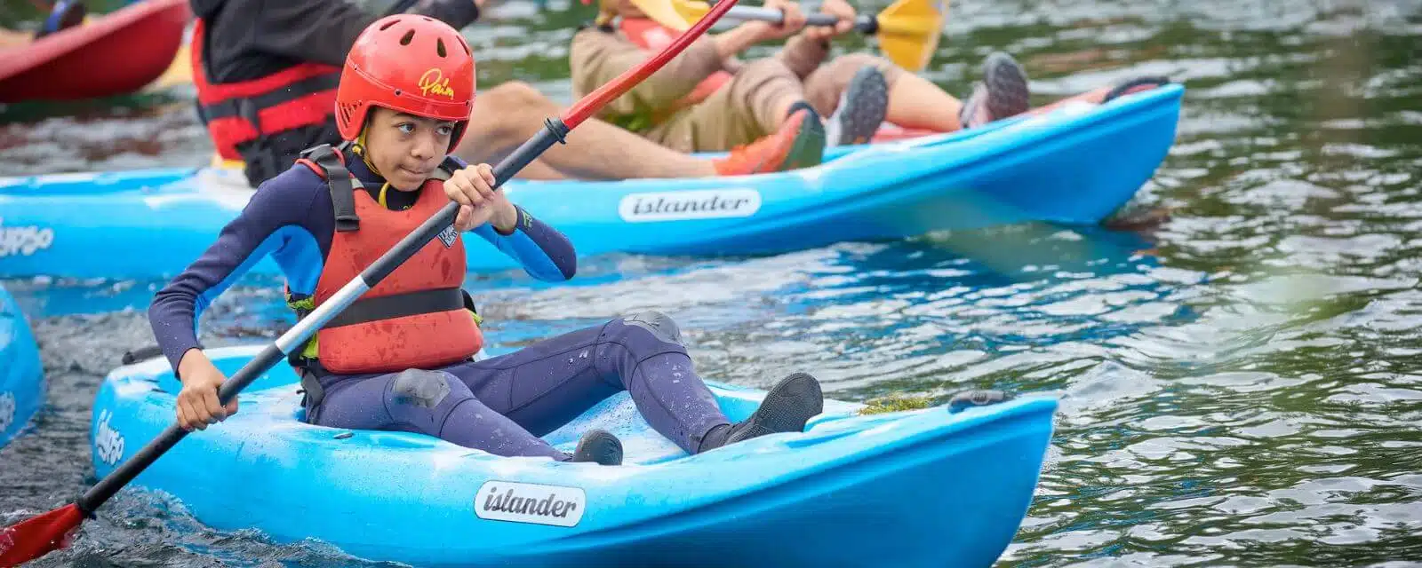 Several people in kayaks paddle on a body of water. The child in the foreground is in a blue kayak and wearing a red helmet and life vest.