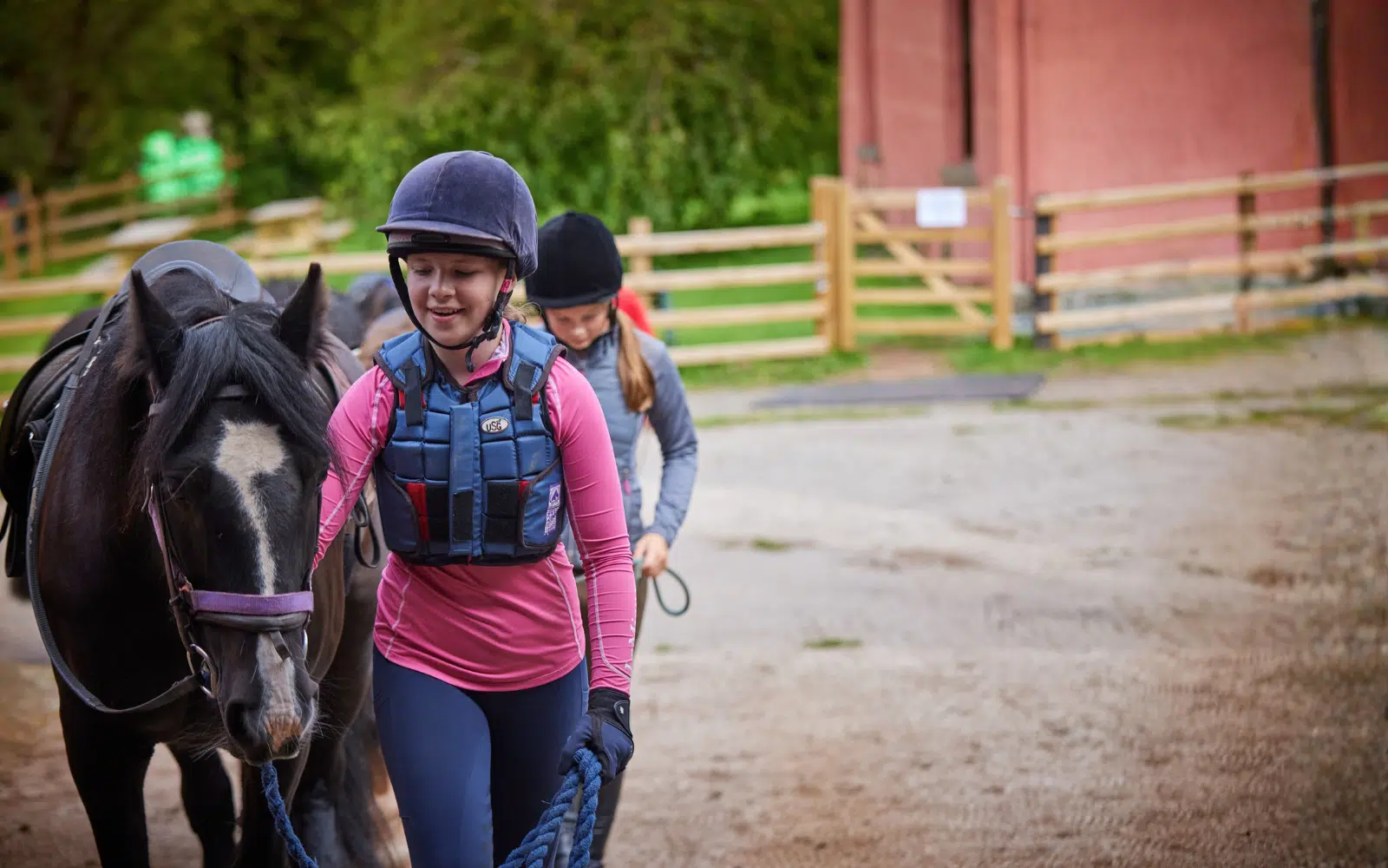Two people wearing helmets and vests lead a black horse with a purple headcollar on a dirt path near a wooden fence and a red building, enjoying an experience reminiscent of PGL Adventure Holidays.
