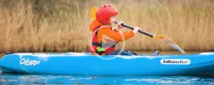A child wearing an orange jacket, red helmet, and life vest paddles a blue kayak labelled "Calypso Islander" on calm water with tall grass in the background, enjoying a fun break from musical theatre camp.