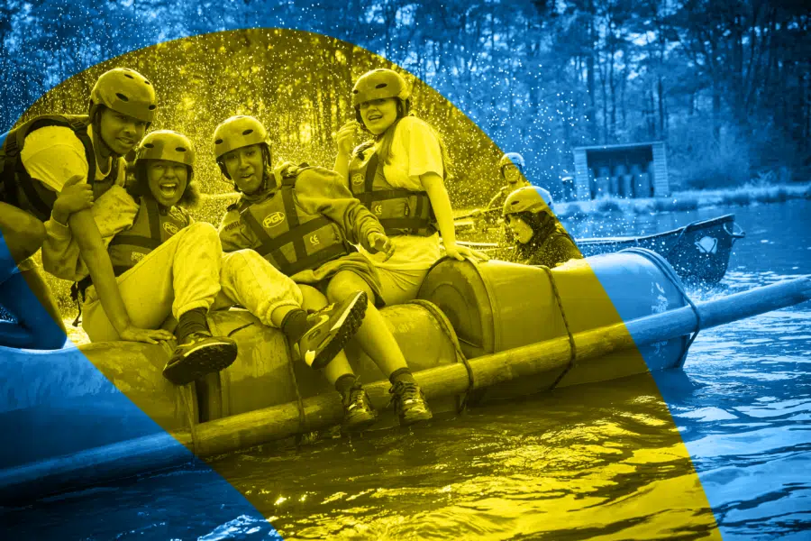 A group of people wearing helmets and life jackets sit on a makeshift raft in a body of water, perhaps enjoying an adventurous team-building activity at musical theatre camp, with trees in the background and water splashing around them.