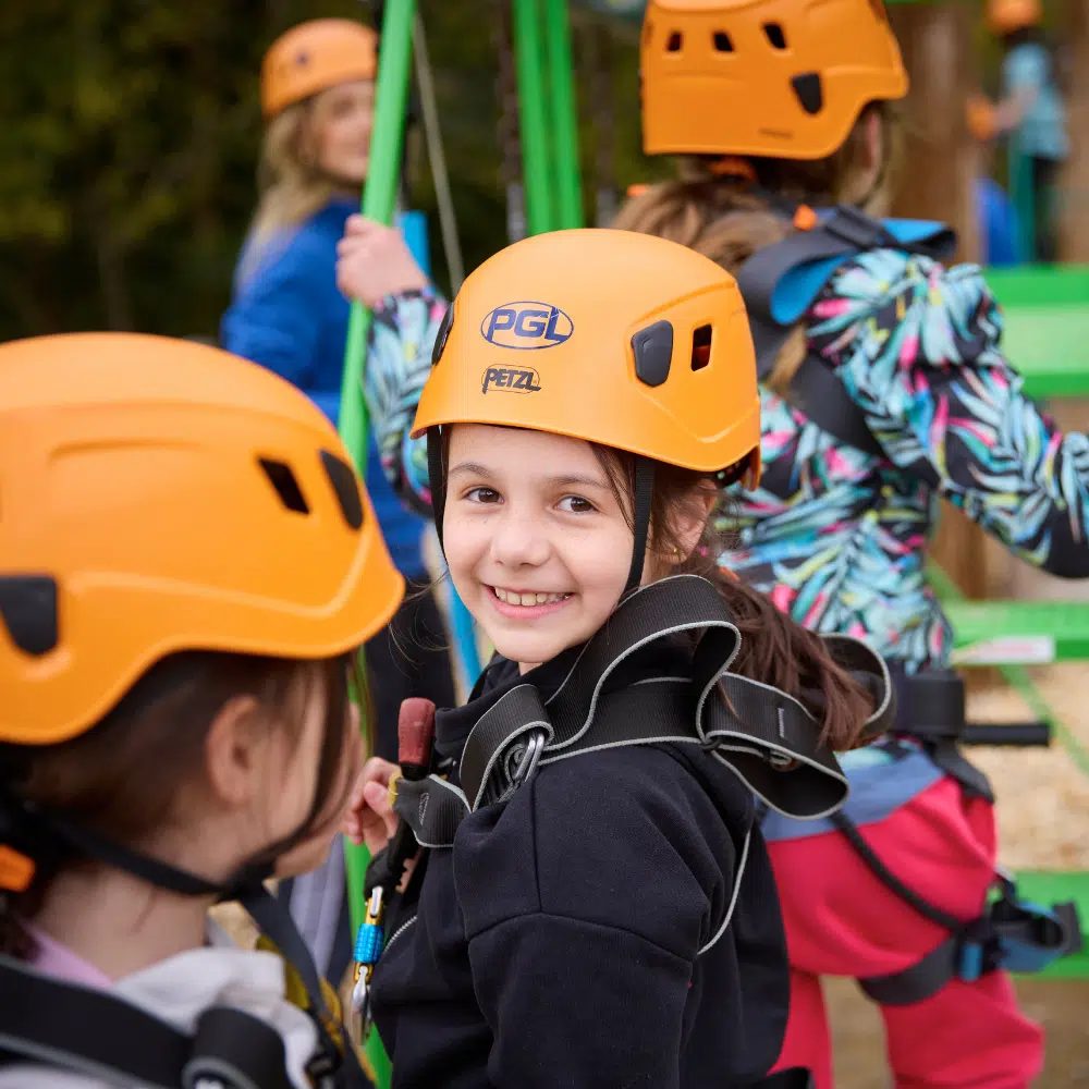 A group of children wearing orange helmets and harnesses stands outdoors. One girl in the centre smiles at the camera whilst others face away.