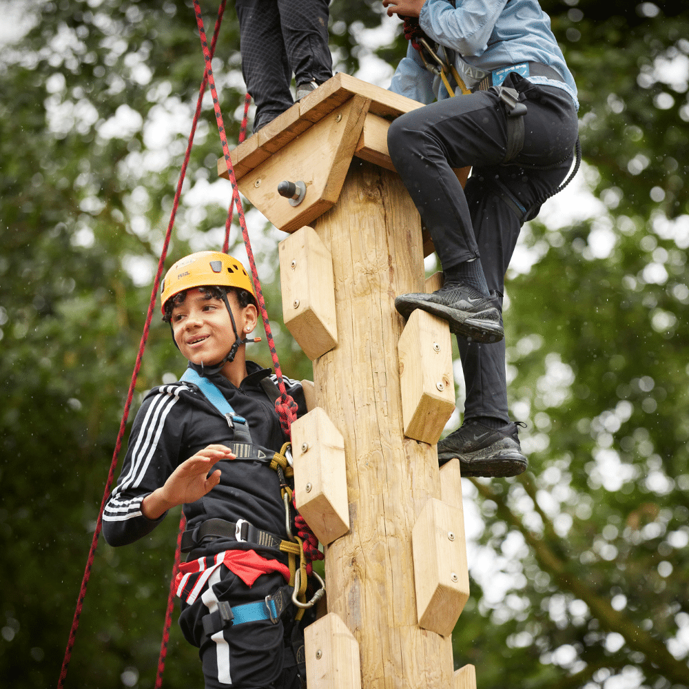 Two people wearing safety gear climb a wooden outdoor adventure pole, surrounded by trees, with one person smiling and looking away from the camera.