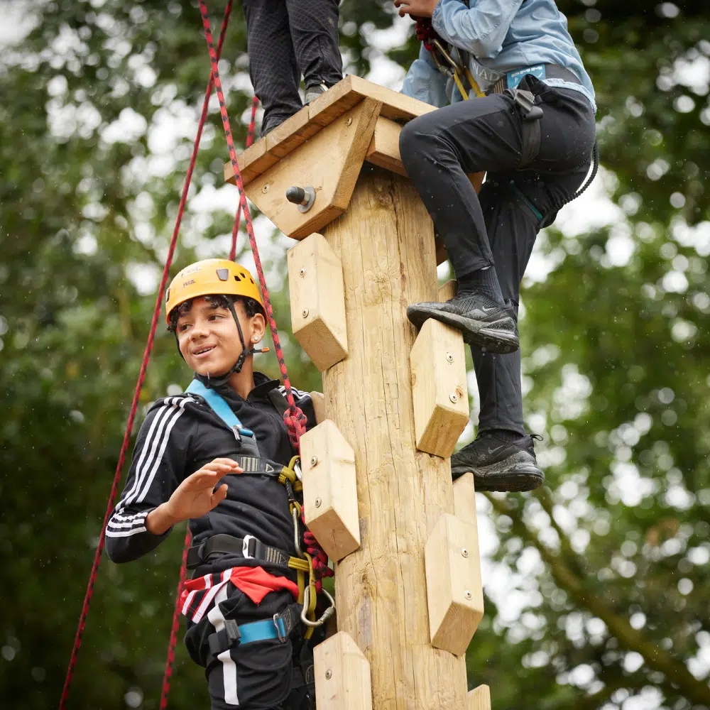 Two people wearing safety gear climb a wooden outdoor adventure pole, surrounded by trees, with one person smiling and looking away from the camera.