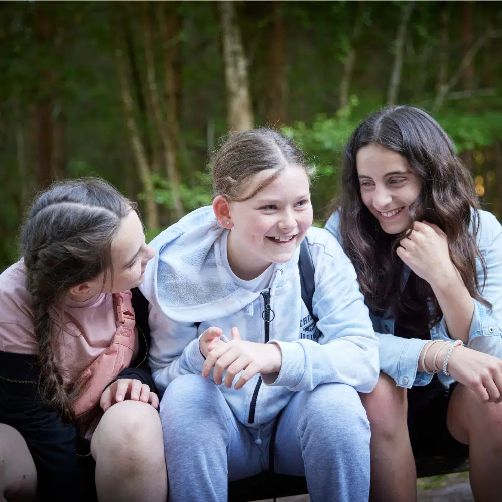 Three girls sit close together outdoors, smiling and talking, with trees in the background.