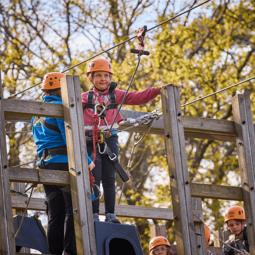 Children wearing helmets and harnesses stand on a wooden platform at an outdoor adventure park, preparing for a zip wire activity. Trees are visible in the background.