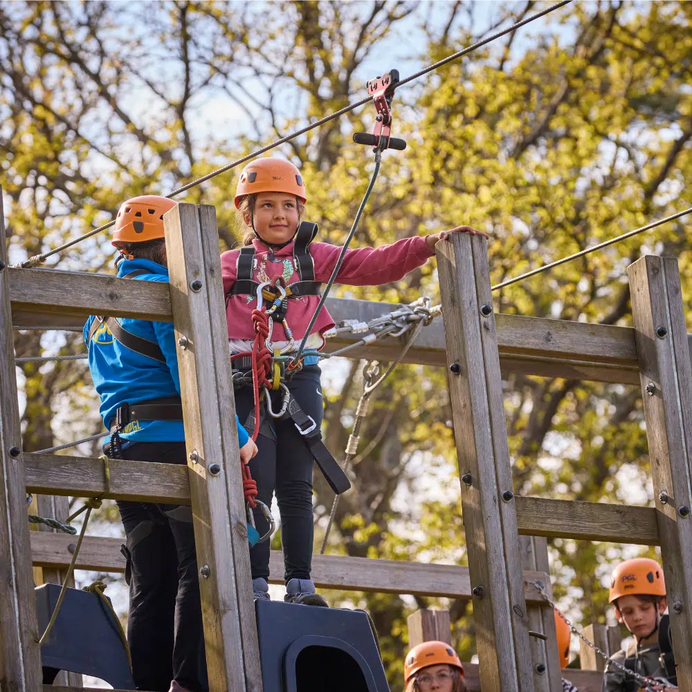 Children wearing helmets and harnesses stand on a wooden platform at an outdoor adventure park, preparing for a zip wire activity. Trees are visible in the background.