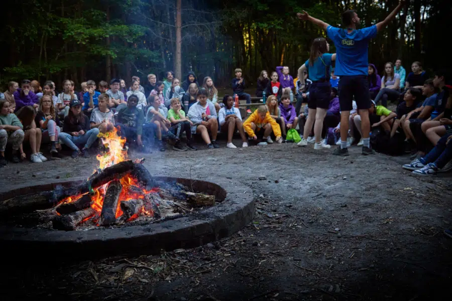 A group of children and adults sit in a circle around a campfire in a wooded area whilst one person stands with arms raised, appearing to lead an activity.