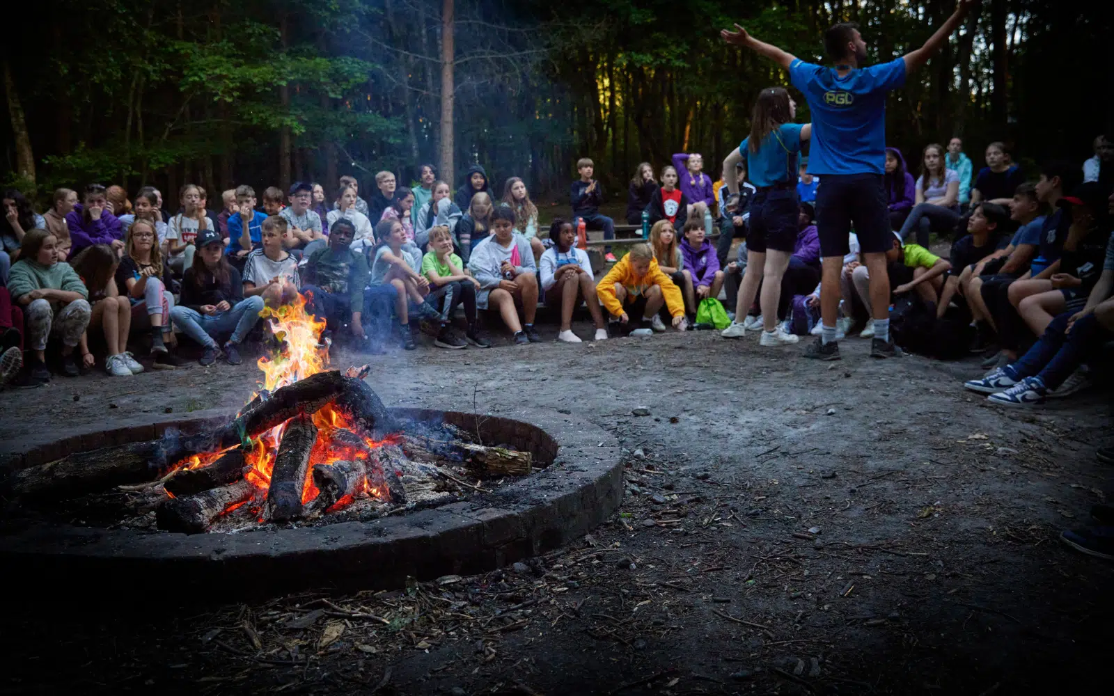 A group of children and adults sit in a circle around a campfire in a wooded area whilst one person stands with arms raised, appearing to lead an activity.
