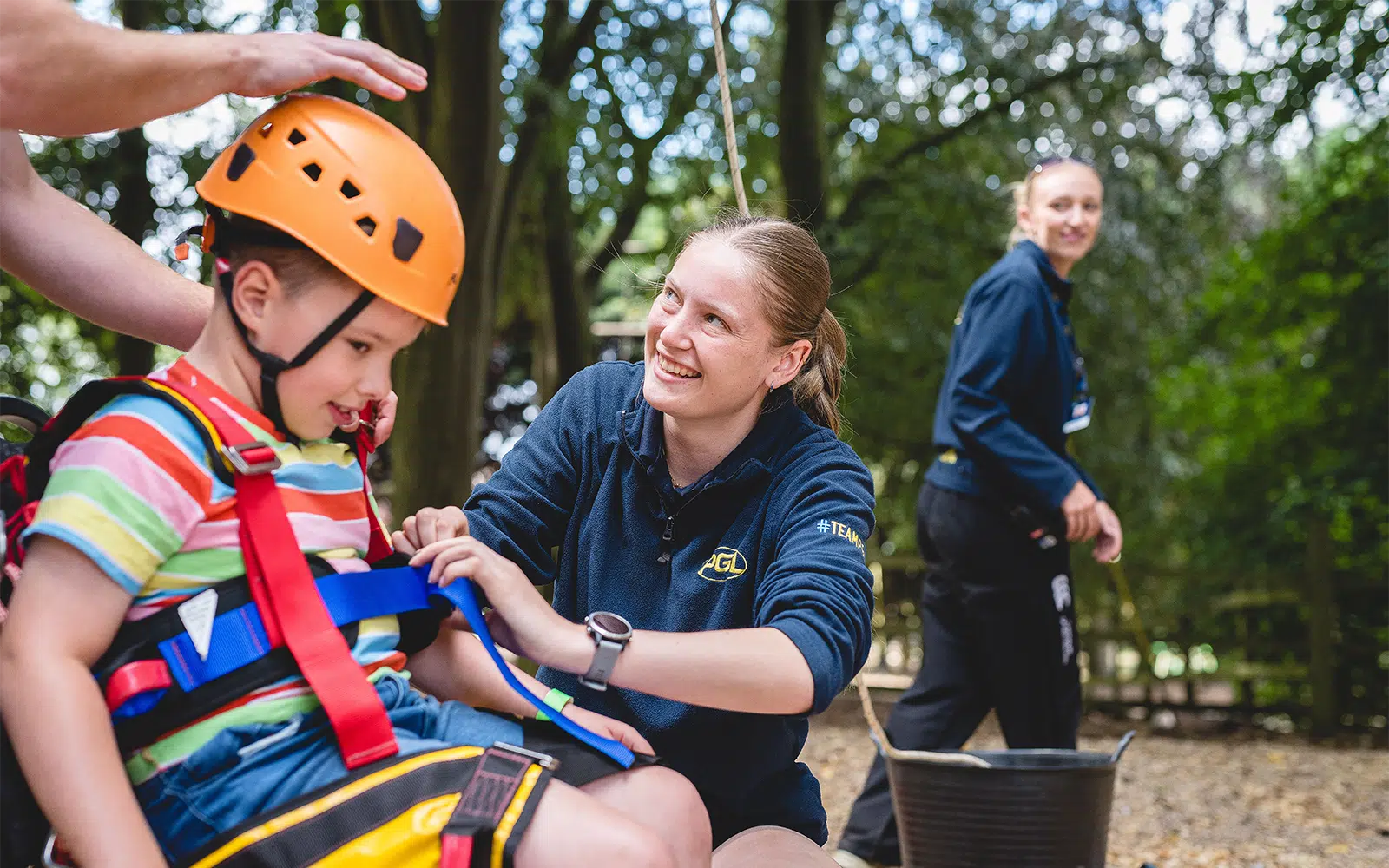 An adult helps a child adjust a safety harness and helmet outdoors, whilst another person stands nearby in a wooded area.