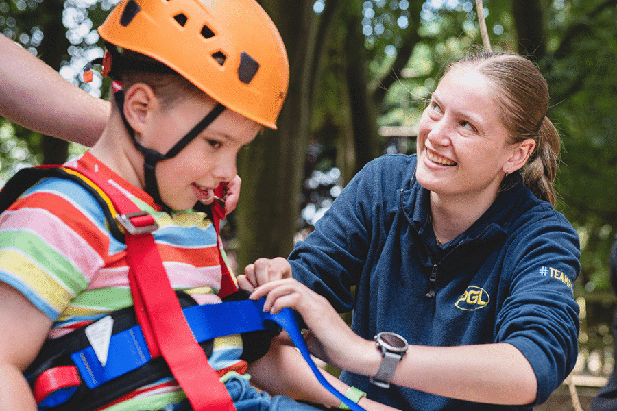 An adult secures a harness on a young child wearing an orange helmet outdoors, whilst another adult stands nearby.