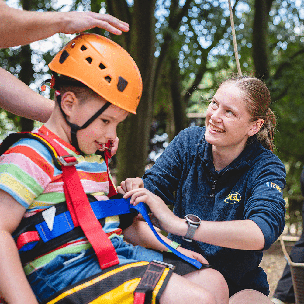 An adult secures a harness on a young child wearing an orange helmet outdoors, whilst another adult stands nearby.