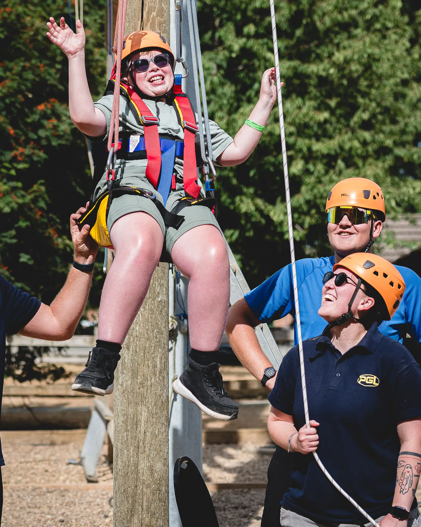 A child wearing a helmet and harness is lifted in the air by ropes, assisted by three adults in safety gear at an outdoor activity centre.