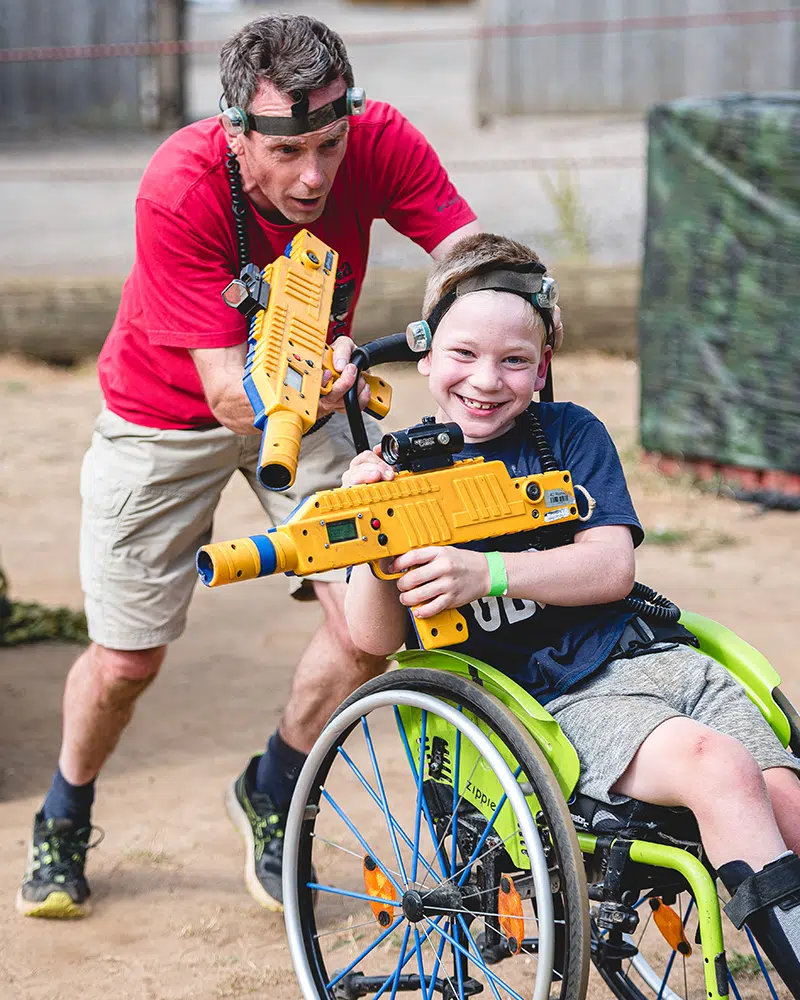 An adult and a child in a wheelchair, both wearing headbands and holding toy laser guns, play together outdoors on a sandy surface.