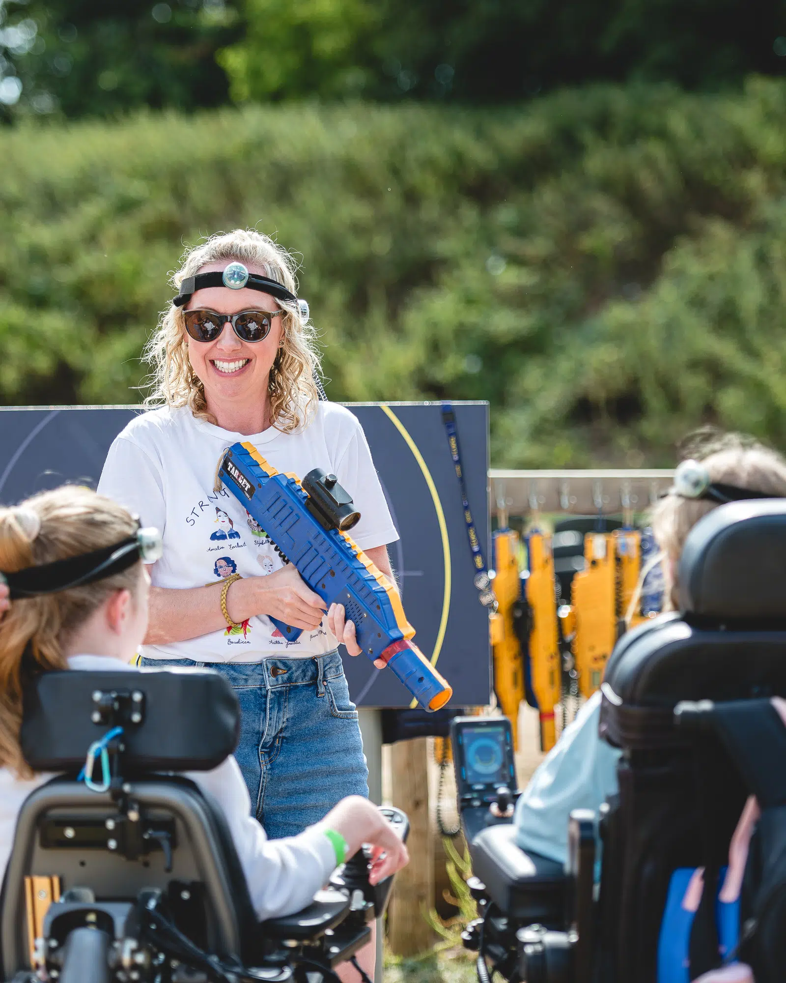 A woman holding a toy blaster talks to two people in wheelchairs outdoors; laser tag equipment is visible in the background.
