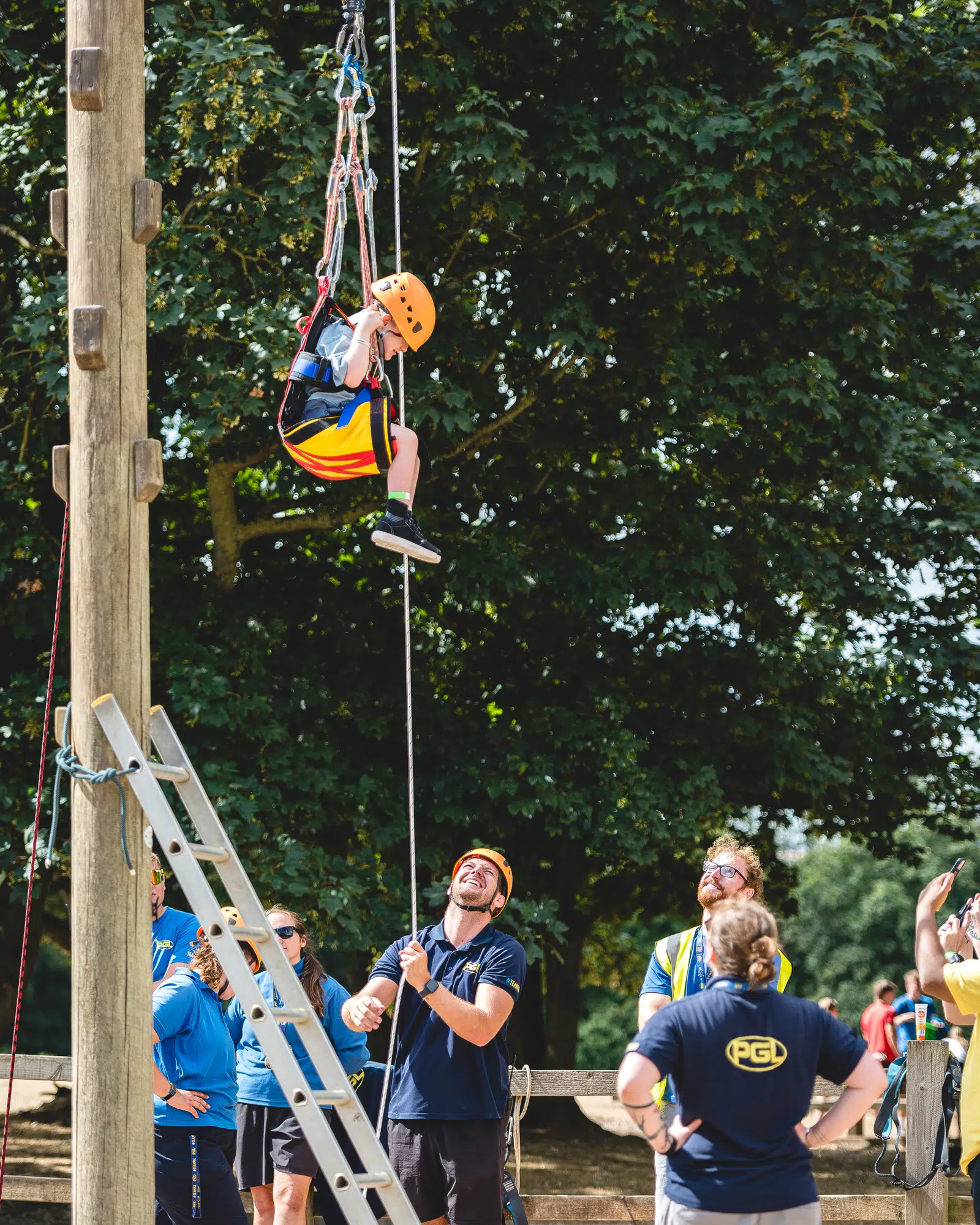 A child wearing a helmet is harnessed and lifted on a ropes course whilst adults watch and guide on the ground.