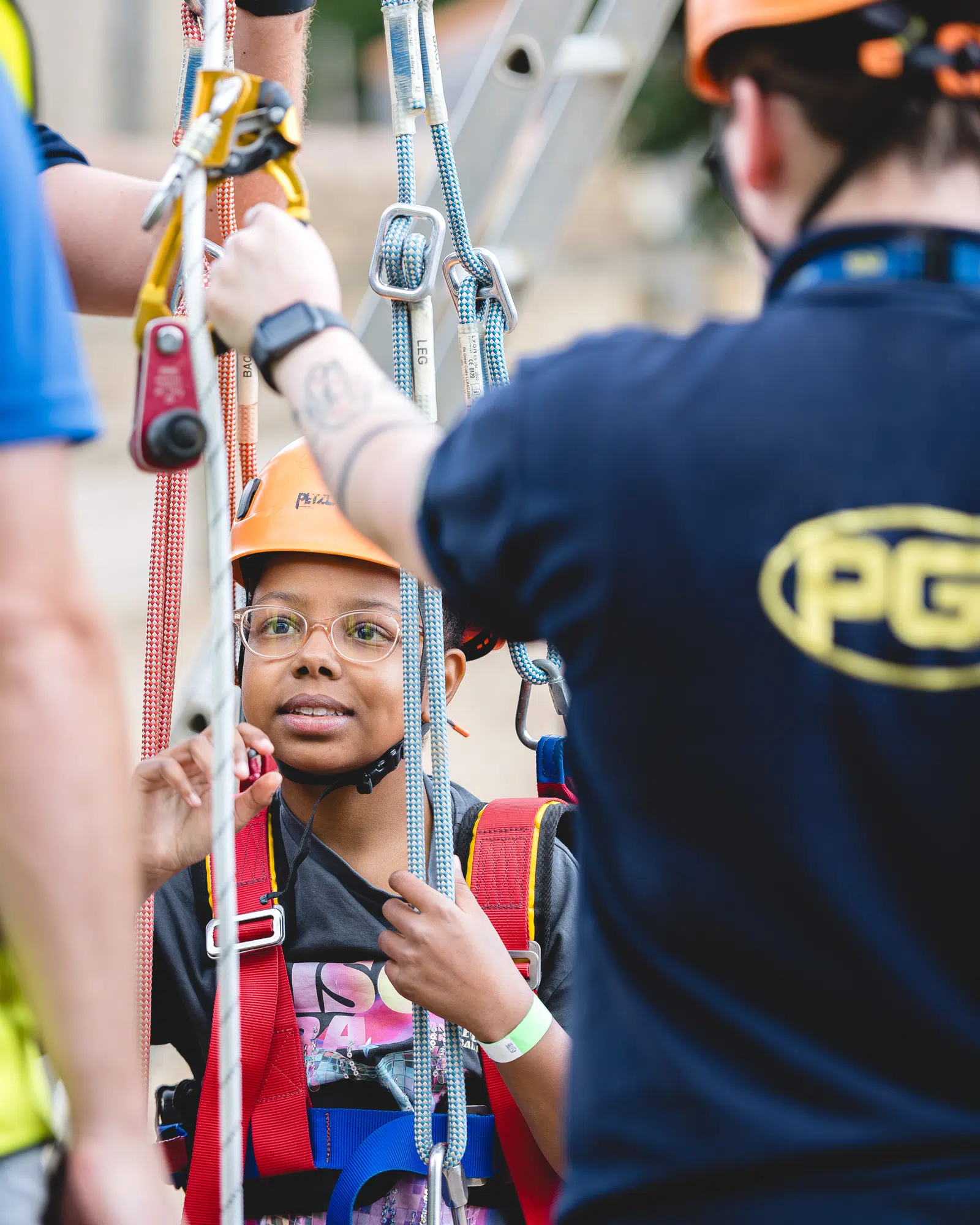 A child in a helmet and harness listens to instructors whilst preparing for a climbing activity. Multiple ropes and safety gear are visible.