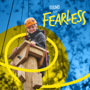 Smiling child in a helmet and harness climbs a wooden pole outdoors, with text reading “Found My Fearless” over a blue and yellow background.