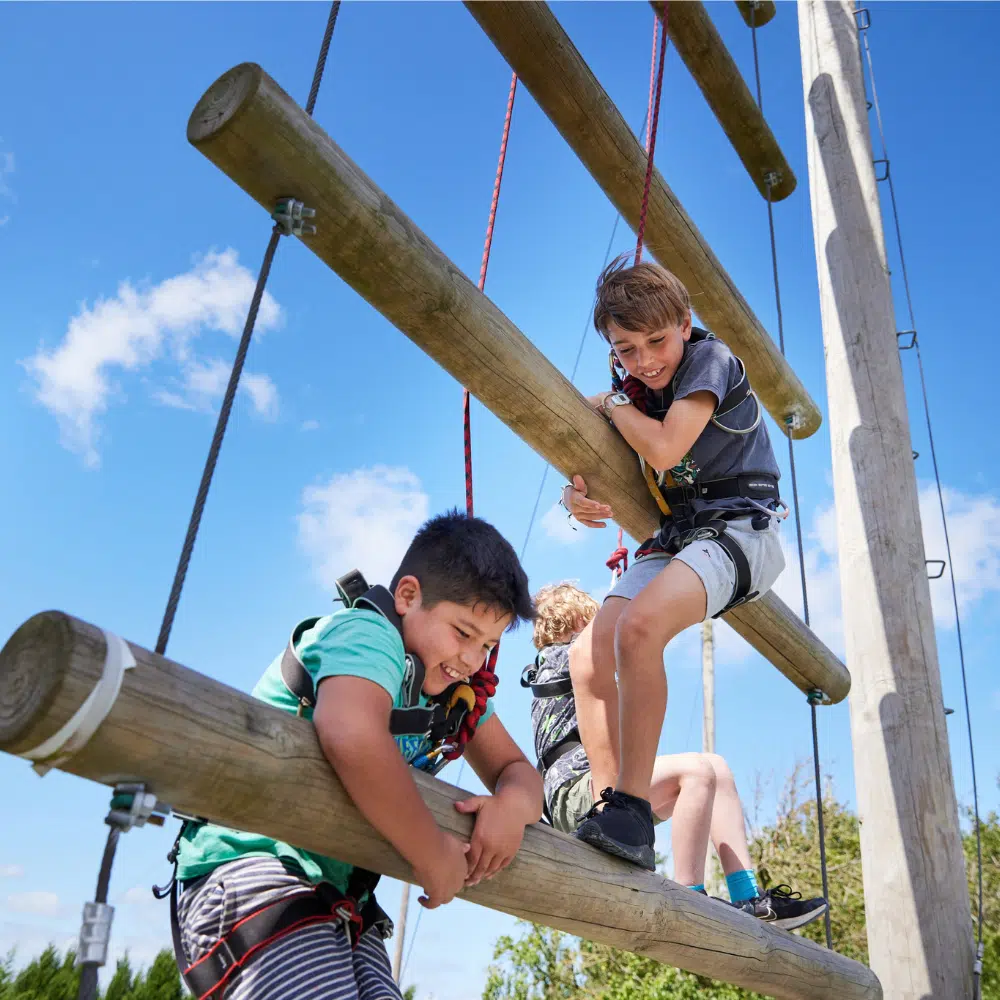 Two children in climbing harnesses navigate a high ropes course made of wooden beams.